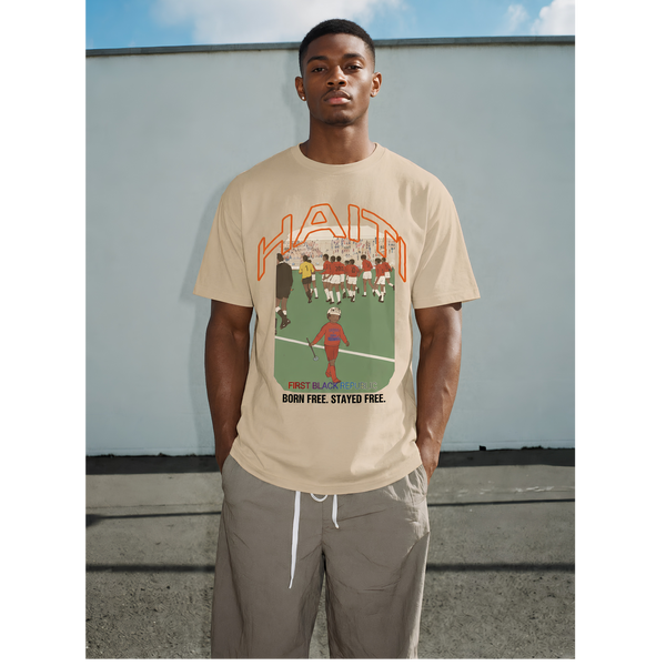 Black male model wearing a tan Haiti graphic T-shirt with “Born Free. Stayed Free.” text, styled with loose pants against a gray outdoor wall.