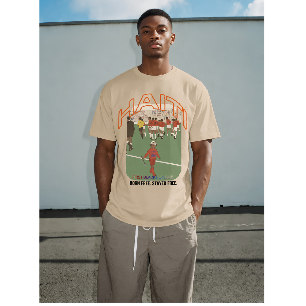 Black male model wearing a tan Haiti graphic T-shirt with “Born Free. Stayed Free.” text, styled with loose pants against a gray outdoor wall. — Breakin Atoms premium streetwear