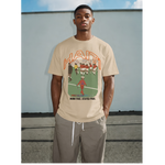 Black male model wearing a tan Haiti graphic T-shirt with “Born Free. Stayed Free.” text, styled with loose pants against a gray outdoor wall. thumbnail 1