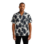 Model wearing Breakin' Atoms "Monochrome Palm Shadow Button-Up," a short sleeve tropical shirt with all-over black palm tree silhouettes on a white background — perfect for vacation or summer streetwear. thumbnail 1