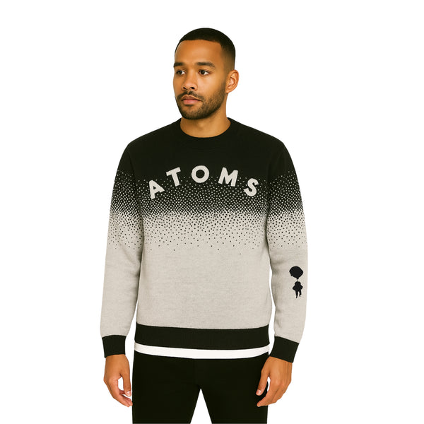 Model wearing Breakin' Atoms "Pixel Dusk Knit," a black-to-gray gradient sweater with pixelated dot fade, bold "ATOMS" typography, and parachuting silhouette graphic on the sleeve — blending cozy knitwear with streetwear flair.