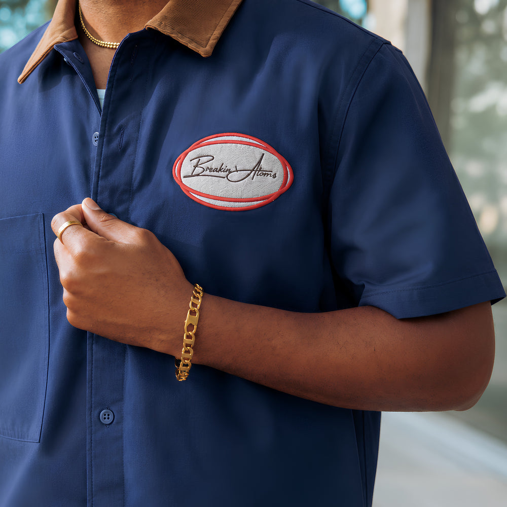 Close-up of a man wearing the Breakin' Atoms Sunday Shift Work Shirt in navy, showing the tobacco corduroy collar, chest pocket, and red-and-grey oval embroidered "Breakin' Atoms" patch, styled with gold jewelry. — Breakin Atoms premium streetwear